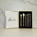 A 5-piece makeup brush set with rose quartz handles, gold ferrules, and white bristles, displayed in a black box next to its white lid which features the brand name 'Arius' in gold foil, set against a white marble background.