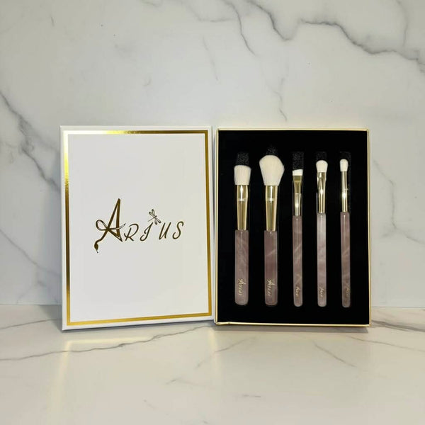 A 5-piece makeup brush set with rose quartz handles, gold ferrules, and white bristles, displayed in a black box next to its white lid which features the brand name 'Arius' in gold foil, set against a white marble background.
